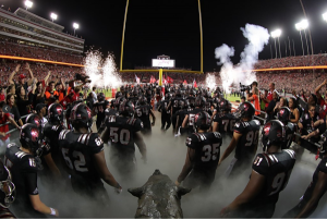 The bronze "Fury" wolf faces the goalpost, and NC State fans cheer as the Wolfpack football team enters the stadium amid smoke and fireworks.